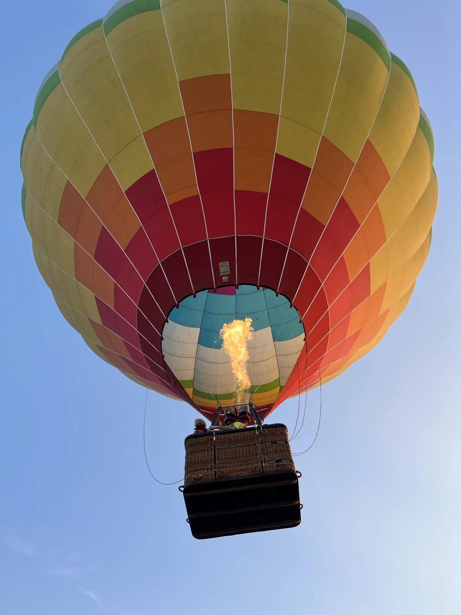 A hot-air balloon in red, orange and yellow seen from below, with its burner lit, against a blue sky